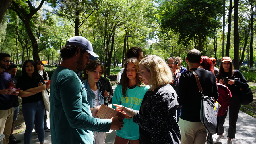 Amid trees, Caitlin Mueller discusses and points to a clay block that someone holds before her, while others look on