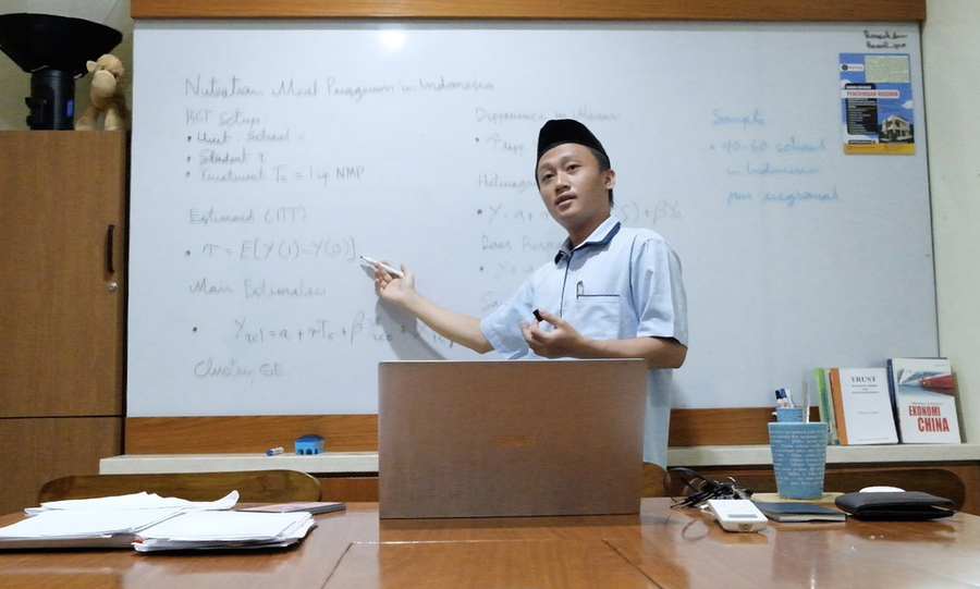 Munip Utama stands before a whiteboard, gesturing. A laptop sits open on the table in front of him.