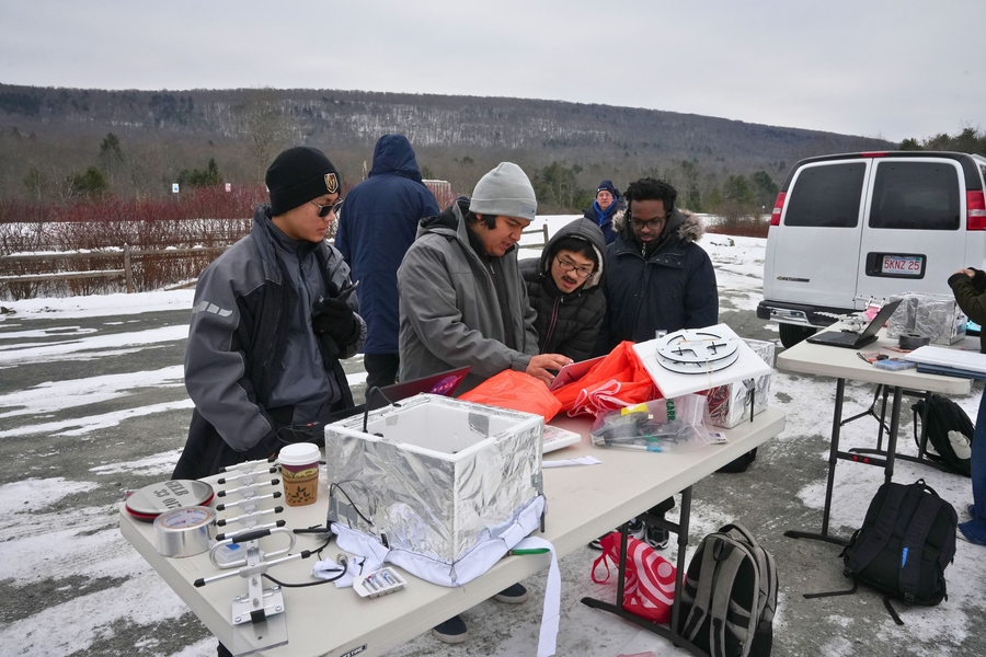 Four students in winter gear stand at a table outdoors next to a van. There is snow on the ground and it's a cloudy day.