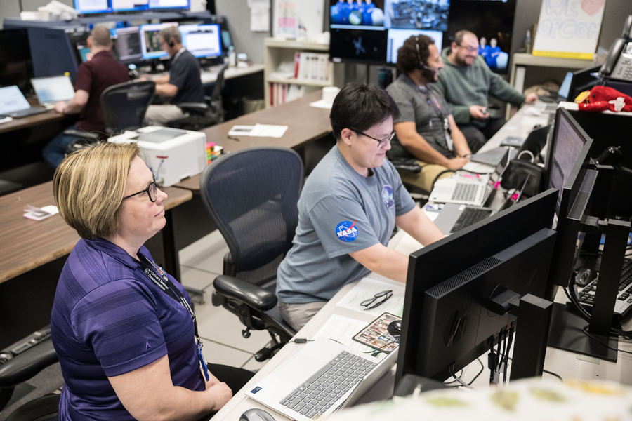 Olga Mikulina and Jesse Chang in the foreground and four people in the background sit in front of computer screens at NASA Mission Control.