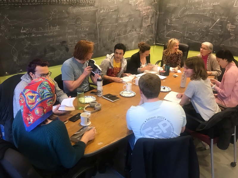10 people sit around a small seminar table with a blackboard in the background