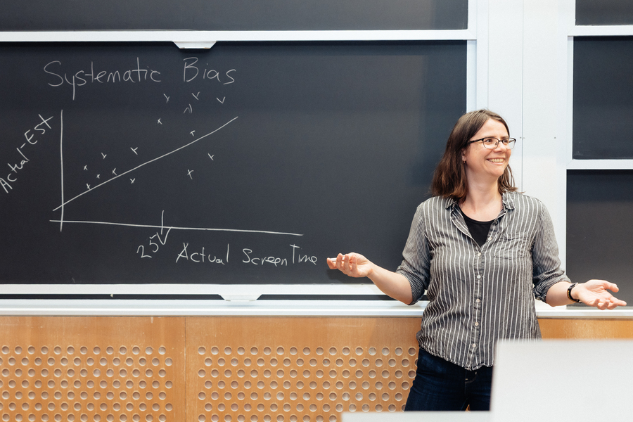Ana Bell stands in front of a blackboard on which she's drawn a simple line graph of actual screentime usage versus estimated screentime usage. The plot shows that many underestimate their screentime usage.