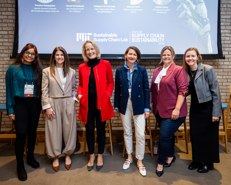 Six women pose standing side by side for a photo before a large display screen indoors.