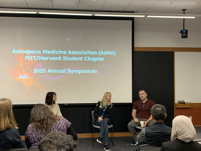 Three people sit facing a classroom, with a projection screen behind them.