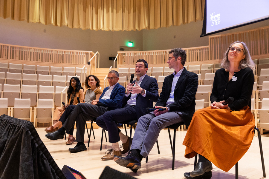 Six people sit in a line in an auditorium with a bank of empty seats behind them. They hold wireless microphones, and one is speaking.