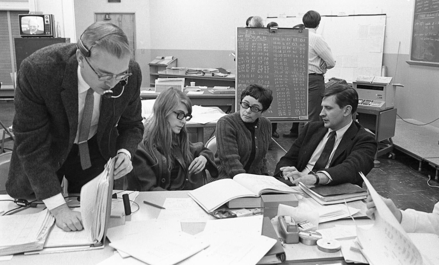 Grayscale photo of Norman Sears, Margaret Hamilton, Phyllis Rye, and Ain Laats conversing at a busy desk in an MIT office in the 1960s.