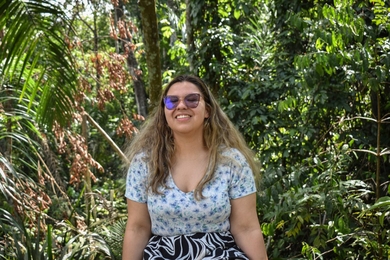 Hana Sousa sits in front of green trees in a rainforest on a sunny day 