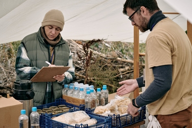 Two people stand outside under a tent. They are taking stock of supplies of bottled water, cans of food, and food packages in crates.