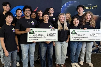 13 CERBERUZ team members pose at a NASA event, smiling and holding oversized $500,000 and $275,000 award checks against a space-themed backdrop.
