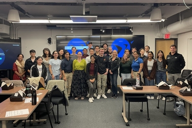 27 students and panelists pose standing in the front of a classroom