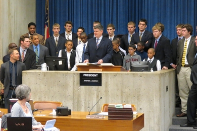 About 17 members of MIT's Men's Basketball team and middle-school aged student athlete surround podium.