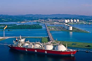Tanker with several large domes floats in a bay where large gas tanks also sit
