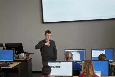 Back view of half a dozen people sitting at long tables, each with a computer monitor in front of them. An instructor is at the front of the class