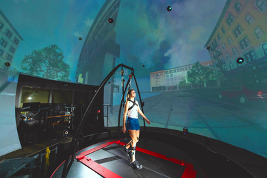Photo of a researcher in a harness walking on a treadmill and surrounded by a spherical screen displaying a city scene.