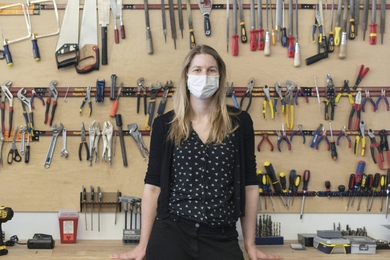 Photo of Stefanie Mueller, masked, standing before a wall holding wrenches, pliars, screwdrivers, saws, and drill bits