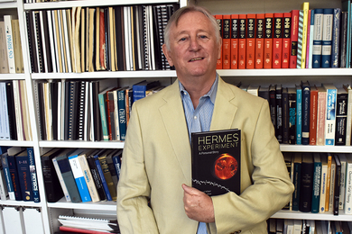 Photo of physics Professor Richard Milner standing in front of a bookshelf, holding a copy of the book, "The Hermes Experiment"
