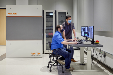 Photo of two researchers looking at images on two computer screens. The VELION FIB-SEM, a large beige cabinet with two racks of electronics, sits behind them.
