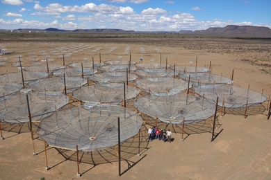 Aerial photo of the HERA radio interferometric array, which consists of many large radio telescopes laid out in a grid in the desert. Eight people stand between two dishes.