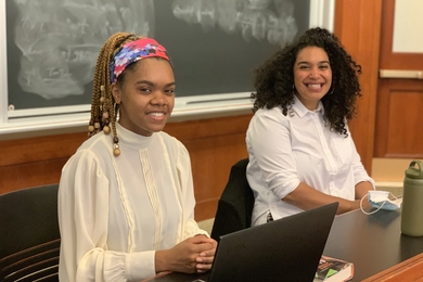 Photo of two women seated before a blackboard, one with a laptop