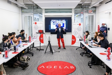 Photo of Joe Coughlin in a bright white-and-red room, standing in front of a screen and addressing students sitting at two long tables on either side of him