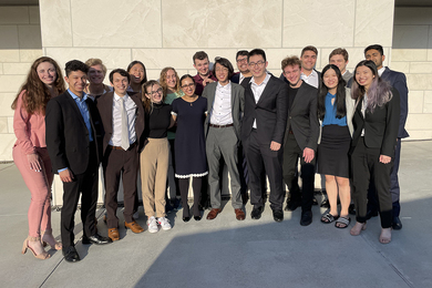Eighteen students stand together posing outside on a sunny day in front of a tan stone wall.