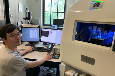Photo of Jiahao Dong sitting at a desk with two monitors in Jaramillo lab. A large piece of lab equipment labeled “TriboIndenter” is in the foreground.