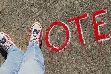 Top-down view of two feet wearing red, white, and blue sneakers positioned in the shape of a V next to letters written in chalk on the ground spelling the word "Vote"