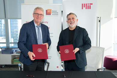 In a conference room, Christoph Meinel and Hashim Sarkis stand in front of banners with Hasso Plattner Institute and MIT logos. Each holds a red folder with the MIT seal on it.