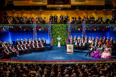 Overview of a large stage with many Nobel Prize winners and guests, all in formal dress, plus the king and queen of Sweden and an orchestra above.