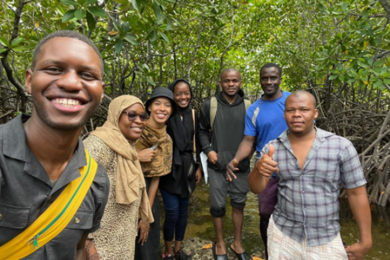 Seven individuals stand and pose for a group photo in a lush green wooded area. 
