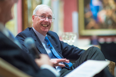 Chris Walsh smiles in a seated position while being interviewed by a blurred figure, seated in the foreground.