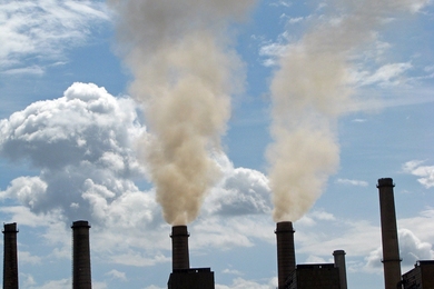 Photo of a row of smokestacks. Two are belching smoke into a cloudy blue sky.