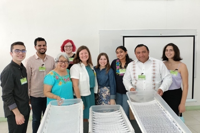 Nine people pose in a line in a white room. Three white prototype bathtubs stand before them