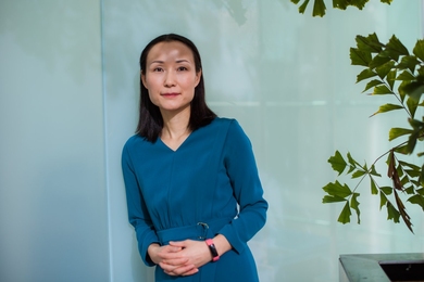Haruko Wainwright leans against an opaque glass wall with a plant in the right side of the frame.