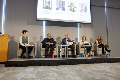 Five people seated in armchairs on a low platform, each with a small round table. A monitor above displays each panelist's name and title with a portrait photo.