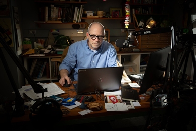 Danny Braunstein, sitting in a pool of light in an otherwise-dim room, looks at a laptop. There is a professional-grade boom microphone in front of him.