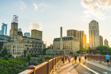 People walk on a pedestrian walkway in front of a city skyline at sunset.