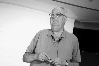 Black-and-white photo of Arnoldo Hax in a polo shirt, standing in front of a blank screen or whiteboard with a microphone clipped to his lapel