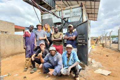 A diverse group of 8 people, five standing and three crouching, pose in front of a shipping container that has been modified for use for cold storage.