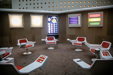 An empty, carpeted room with red-and-white chairs arranged in a circle and screens on the wall creating a futuristic appearance
