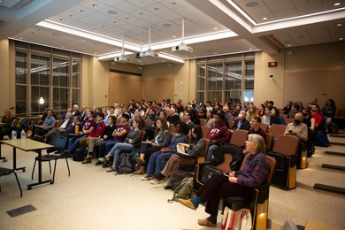In a small auditorium, about 100 people sit listening to a speaker who is out of frame