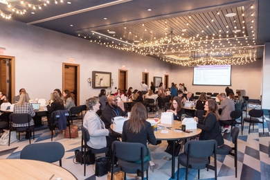 About 40 people are visible eating lunch at eight round tables in a cafeteria. There’s a projection screen on the far wall.