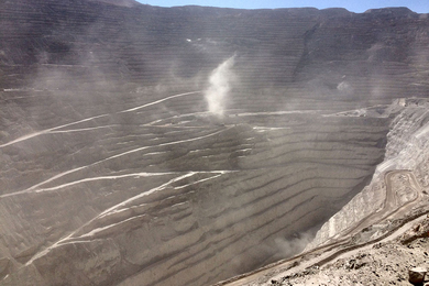 A copper mining pit dug deep in the ground, surrounded by grooved rings of rock and flying dust.