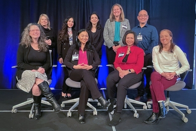 A group photo of eight women and one man in two rows, with back row standing and front seated, on a platform with dark curtains behind them.