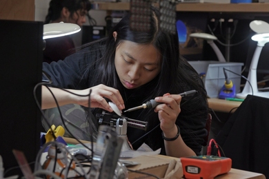 Seated at a lab table, Audrey Chen solders a circuit board