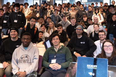 About 100 people pose for a photo in a large classroom. About a third are seated in the foreground and the rest stand