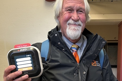David Barber stands in an MIT office and holds an AED device up to the camera. The device’s touchscreen display is illuminated.