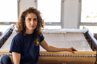 Chloe Bensahel sits in front of a loom, looking at the camera