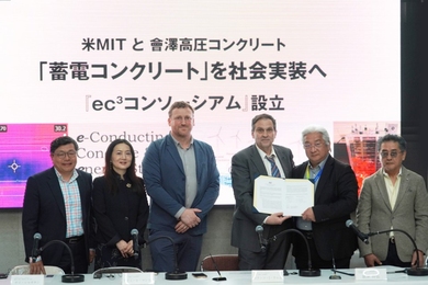 Six people stand behind a long desk with Japanese characters projected behind them. Two men hold a signed agreement.