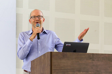 Evan Lieberman, holding a microphone, speaks at a lectern bearing an open laptop